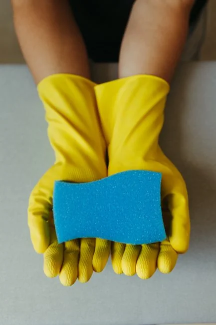 Close-up of hands wearing yellow gloves next to blue sponge on gray surface.